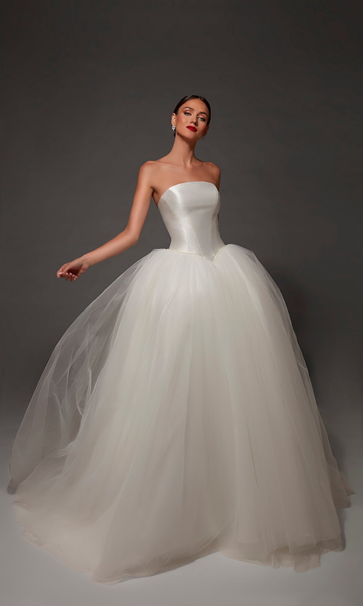 A woman wearing the Long Wedding Dress: Alyce 7182 in Diamond White, featuring a strapless mikado bodice and a voluminous tulle skirt, stands against a dark background.